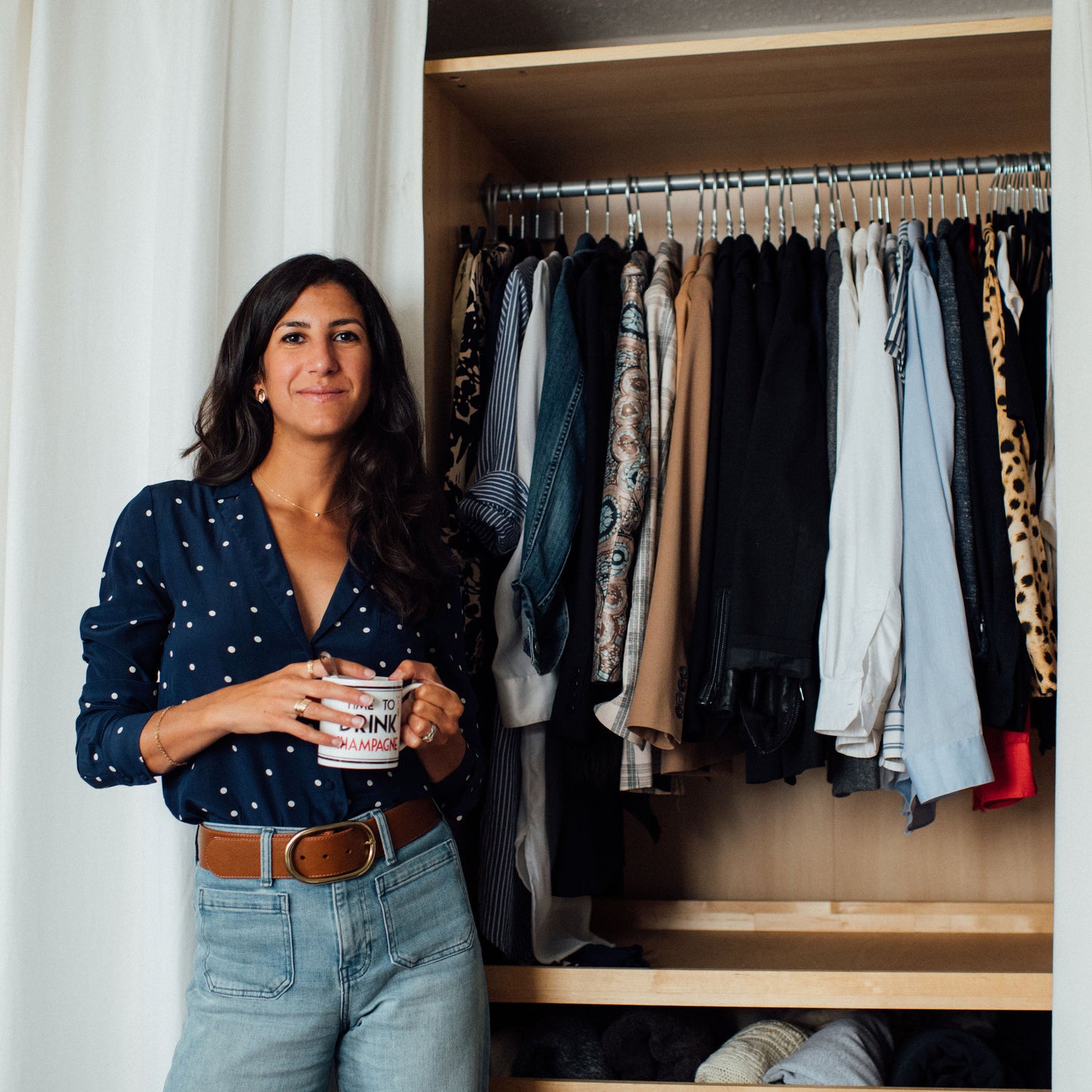 alyssa beltempo is a slow fashion stylist who offers virtual "shop your closet" sessions. She is standing holding coffee next to a neatly arranged closet full of beautiful clothes